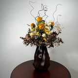 A dried flower arrangement in a dark ribbed vase, displayed on a round wooden table against a plain light gray wall.