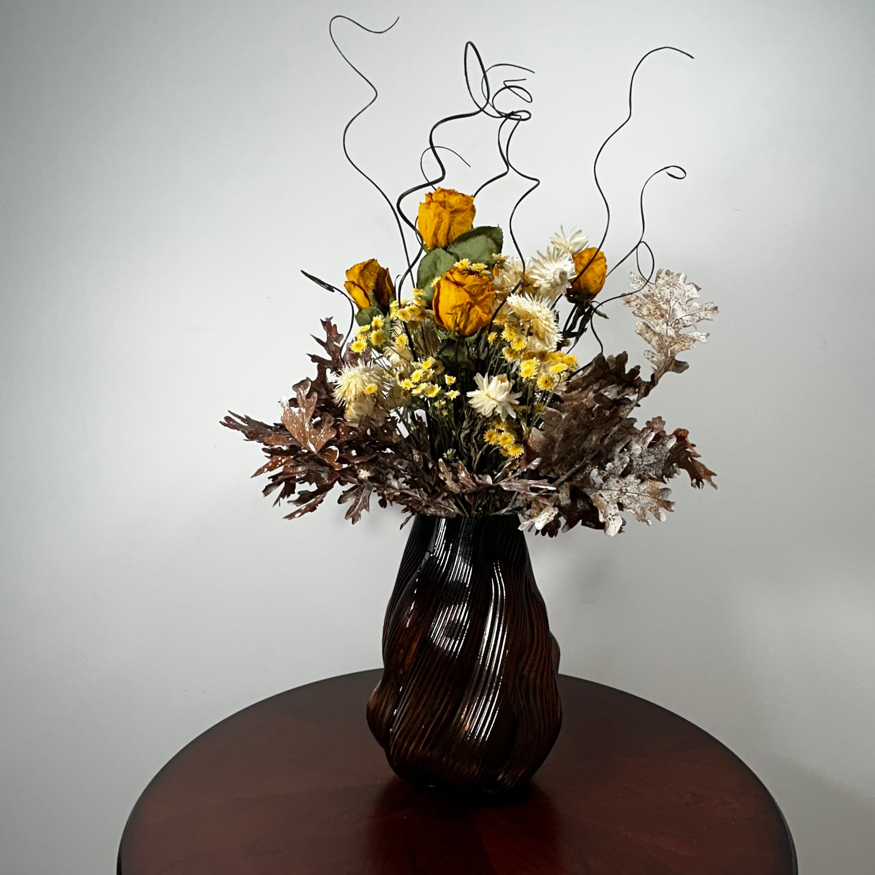 A dried flower arrangement in a dark ribbed vase, displayed on a round wooden table against a plain light gray wall.