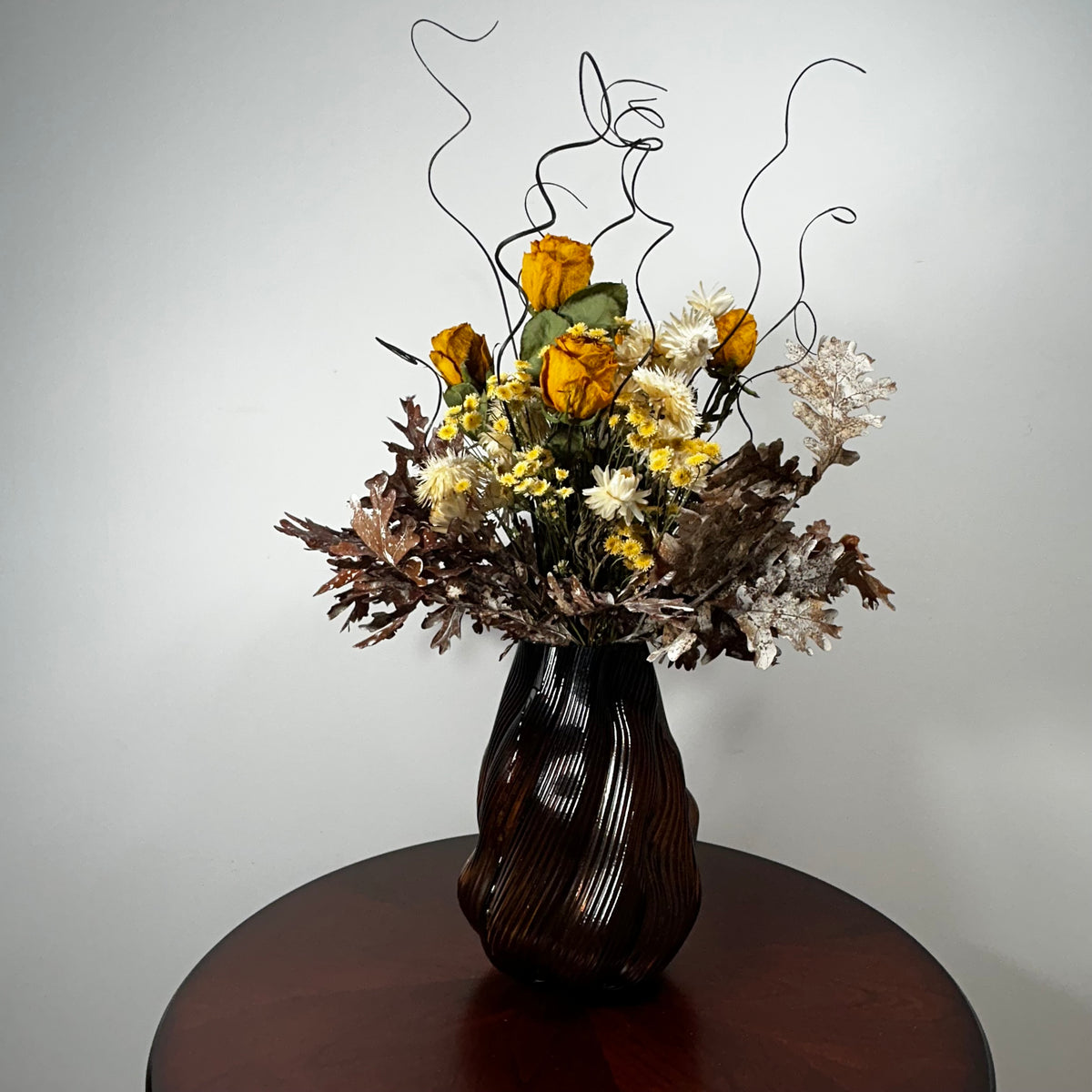 A dried flower arrangement in a dark ribbed vase, displayed on a round wooden table against a plain light gray wall.