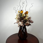 A dried floral arrangement in a dark ribbed ceramic vase, displayed on a round wooden table against a plain light gray wall.