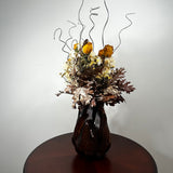 A dried floral arrangement in a dark ribbed ceramic vase, displayed on a round wooden table against a plain light gray wall.