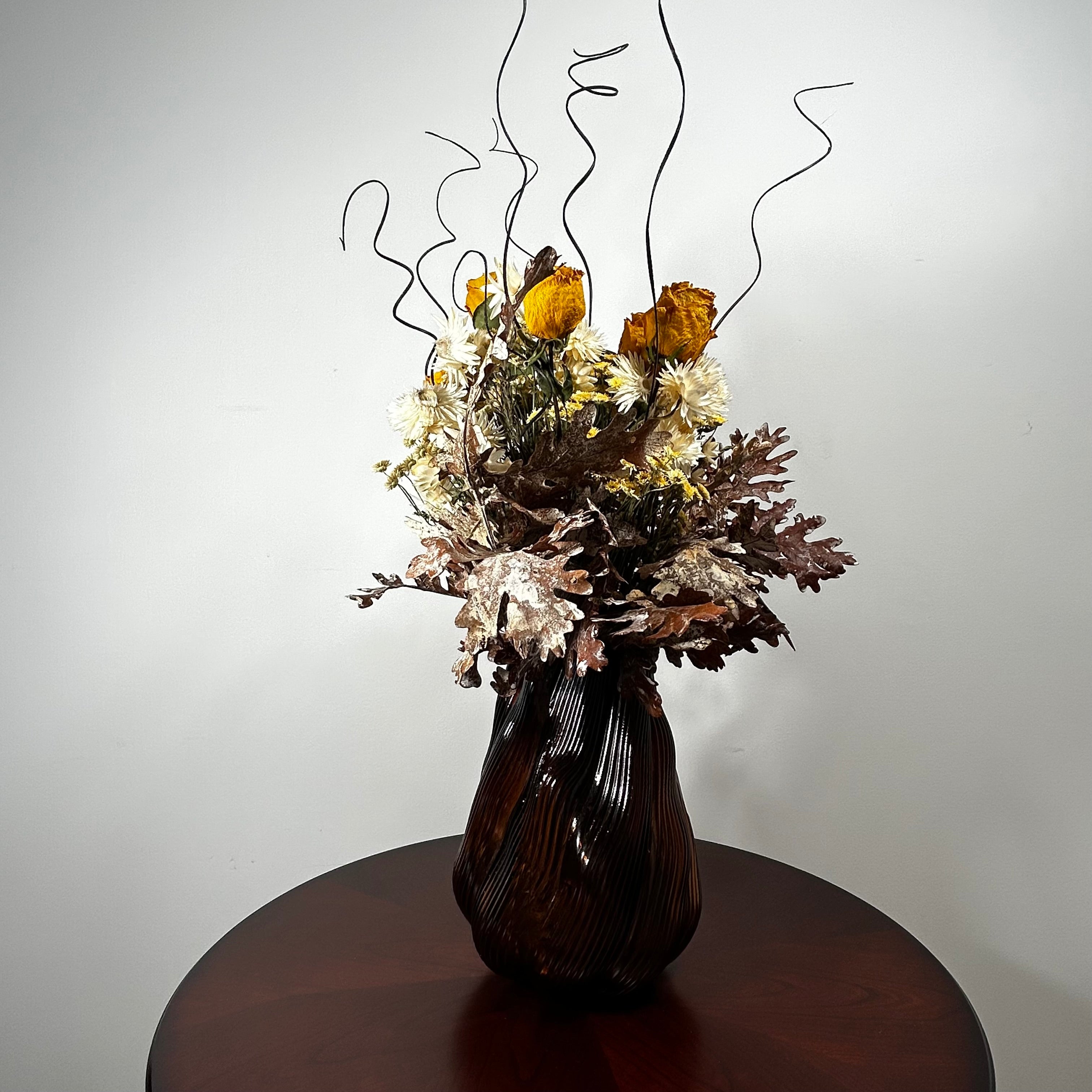 A dried floral arrangement in a dark ribbed ceramic vase, displayed on a round wooden table against a plain light gray wall.
