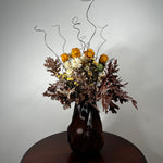 A dried flower arrangement in a dark ribbed glass vase, placed on a round wooden table against a plain light gray wall. The bouquet includes orange roses, white daisies, brown and burgundy foliage, and thin black wire accents rising above the flowers.