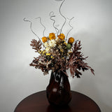 A dried flower arrangement in a dark ribbed glass vase, placed on a round wooden table against a plain light gray wall. The bouquet includes orange roses, white daisies, brown and burgundy foliage, and thin black wire accents rising above the flowers.