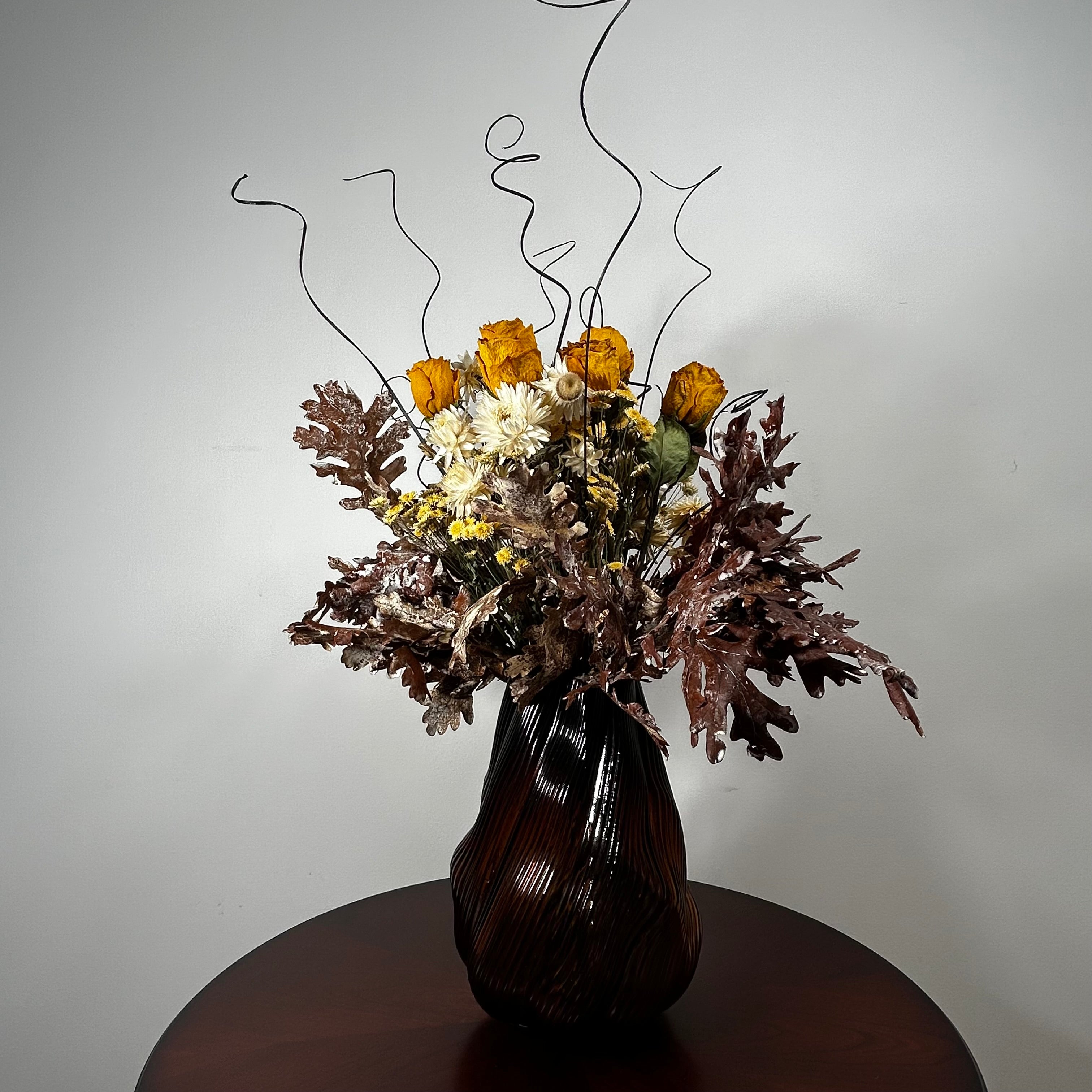A dried flower arrangement in a dark ribbed glass vase, placed on a round wooden table against a plain light gray wall. The bouquet includes orange roses, white daisies, brown and burgundy foliage, and thin black wire accents rising above the flowers.