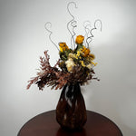 A dried flower arrangement in a dark brown ribbed glass vase, displayed on a round wooden table against a plain light gray wall.