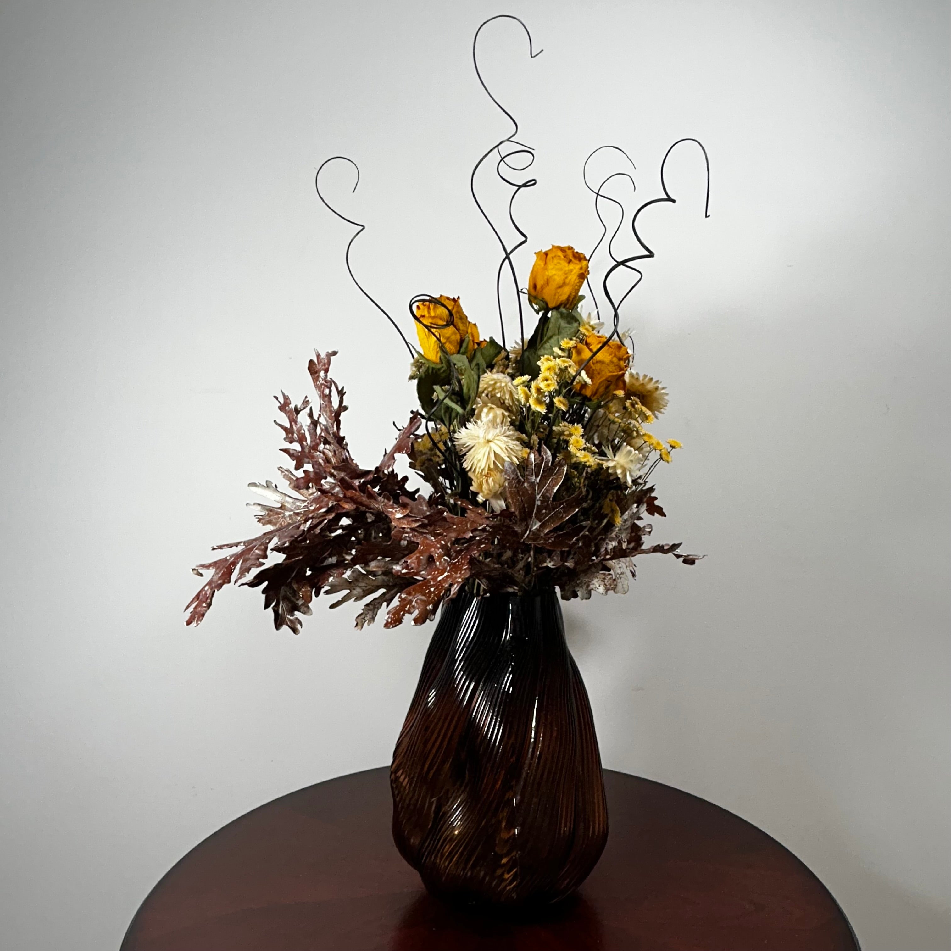 A dried flower arrangement in a dark brown ribbed glass vase, displayed on a round wooden table against a plain light gray wall.