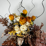 A dried floral arrangement featuring yellow-orange roses, white daisies, small yellow flowers, and textured brown and burgundy leaves, styled with dark wire accents against a plain light gray background.