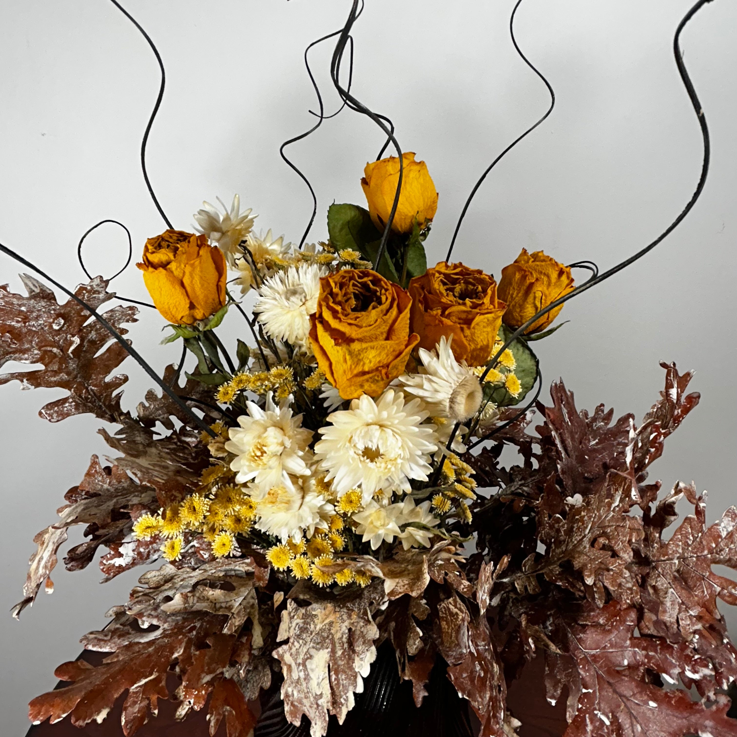 A dried floral arrangement featuring yellow-orange roses, white daisies, small yellow flowers, and textured brown and burgundy leaves, styled with dark wire accents against a plain light gray background.