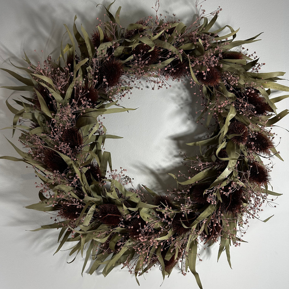 A dried floral wreath featuring olive green leaves, dark brown seed pods, and delicate dusty pink baby's breath, displayed against a plain light gray wall.
