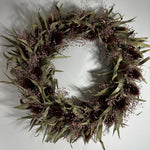 A dried floral wreath featuring olive green leaves, dark brown seed pods, and delicate dusty pink baby's breath, displayed against a plain light gray wall. 1