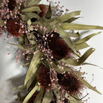A dried floral arrangement featuring dark brown spiky thistle-like blooms, muted olive-green leaves, and delicate pale pink baby's breath stems on a plain light background. 2