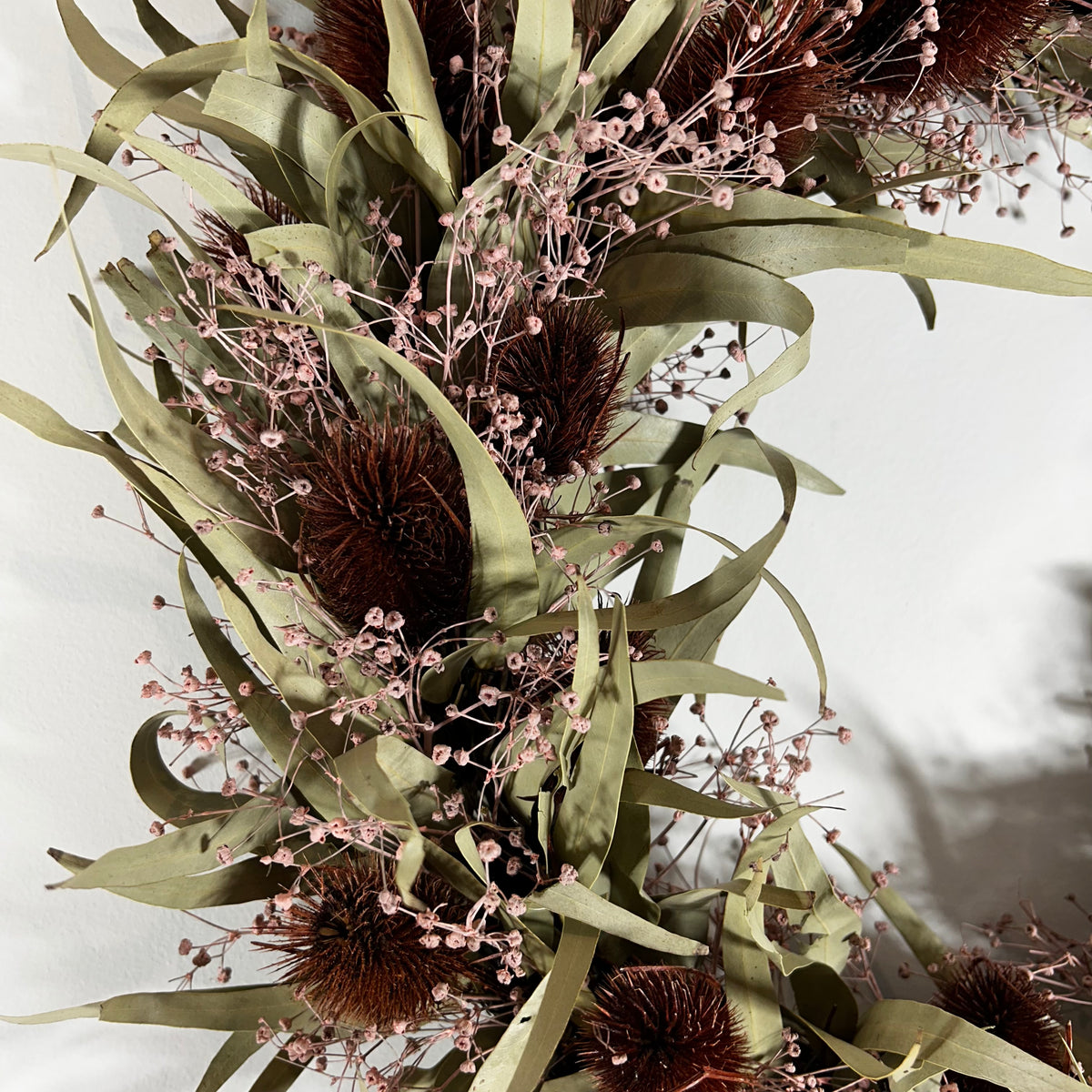 A dried floral arrangement featuring olive-green leaves, dark brown spiky spheres, and delicate pale pink baby's breath on a white background.