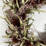 A dried floral arrangement featuring olive-green leaves, dark brown spiky spheres, and delicate pale pink baby's breath on a white background. 3