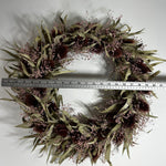 A dried floral wreath made of olive-green leaves, dark brown seed pods, and small pale pink flowers, shown on a white background with a metal ruler across the middle for scale. 4