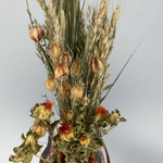Top view of a decorative glass vase shaped like a paper bag with a leather handle filled with dried helichrysum, a beautifully arranged array of natural oats, Nigel, and safflower rise up from the vase.