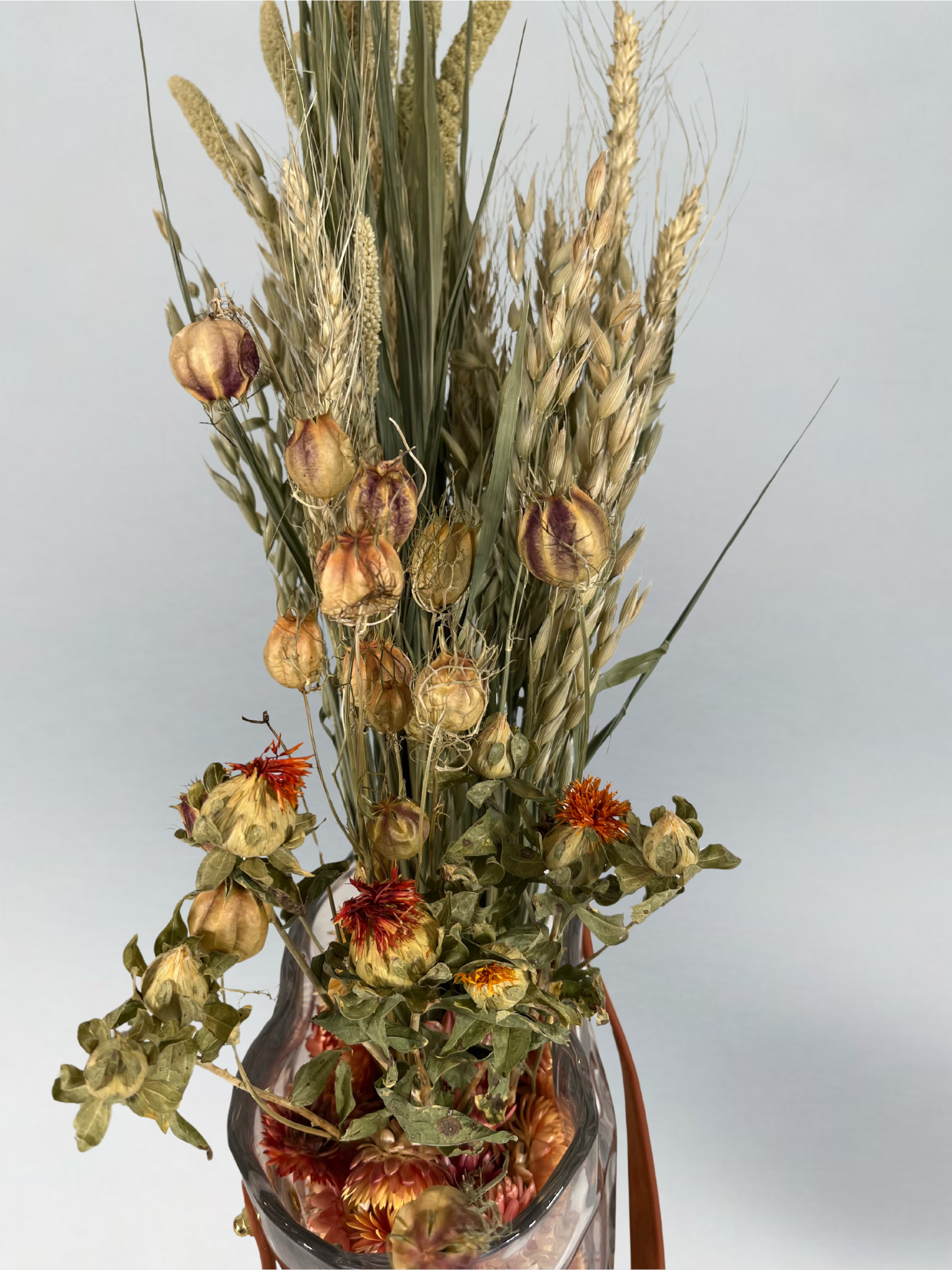 Top view of a decorative glass vase shaped like a paper bag with a leather handle filled with dried helichrysum, a beautifully arranged array of natural oats, Nigel, and safflower rise up from the vase.