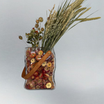 Decorative glass vase shaped like a paper bag with a leather handle filled with dried helichrysum, a beautifully arranged array of natural oats, Nigel, and safflower rise up from the vase.