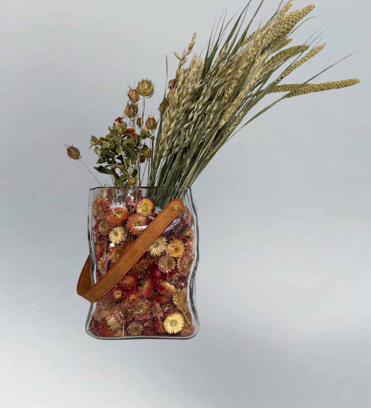 Decorative glass vase shaped like a paper bag with a leather handle filled with dried helichrysum, a beautifully arranged array of natural oats, Nigel, and safflower rise up from the vase.