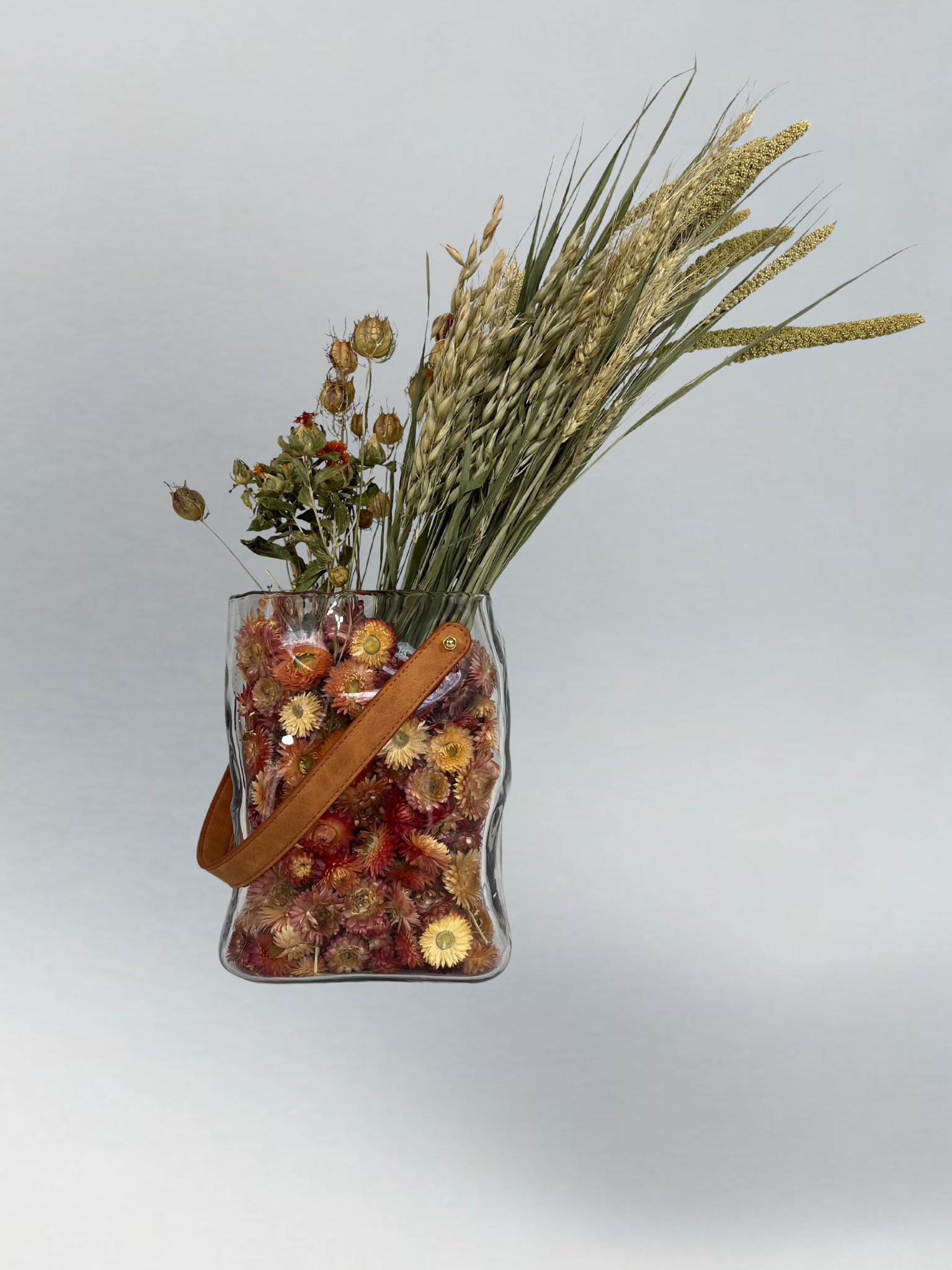 Decorative glass vase shaped like a paper bag with a leather handle filled with dried helichrysum, a beautifully arranged array of natural oats, Nigel, and safflower rise up from the vase.