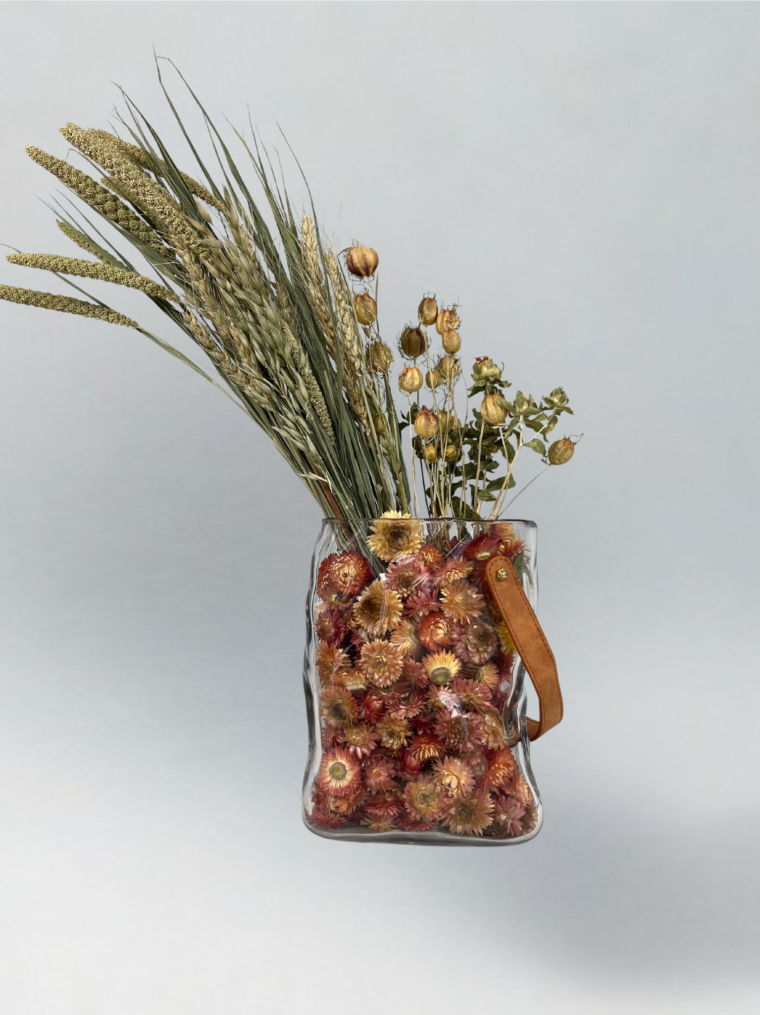 Decorative glass vase shaped like a paper bag with a leather handle filled with dried helichrysum, a beautifully arranged array of natural oats, Nigel, and safflower rise up from the vase.