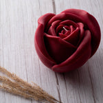 Red rose-shaped candle on a light wooden surface with dried grasses.