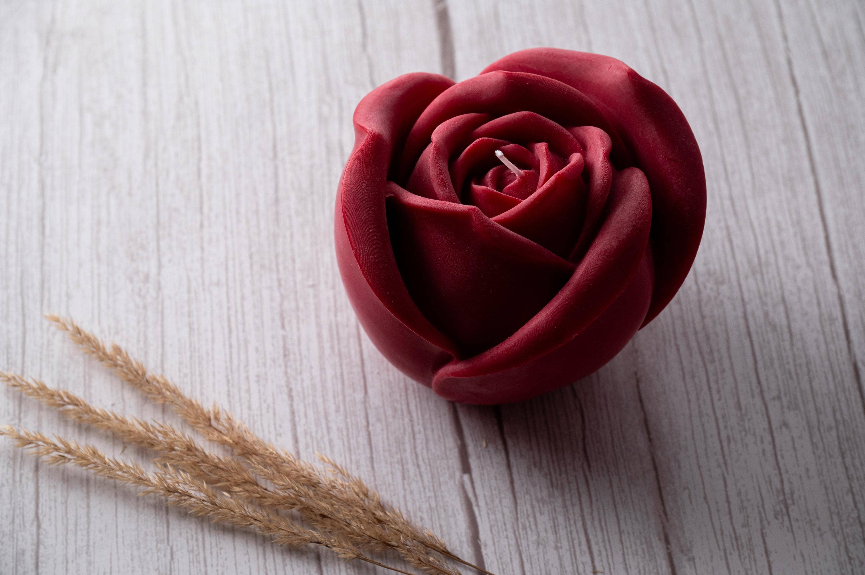 Red rose-shaped candle on a light wooden surface with dried grasses.