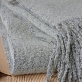 A luxurious wool blend boucle throw in grey with tassels draped over a wicker chair.