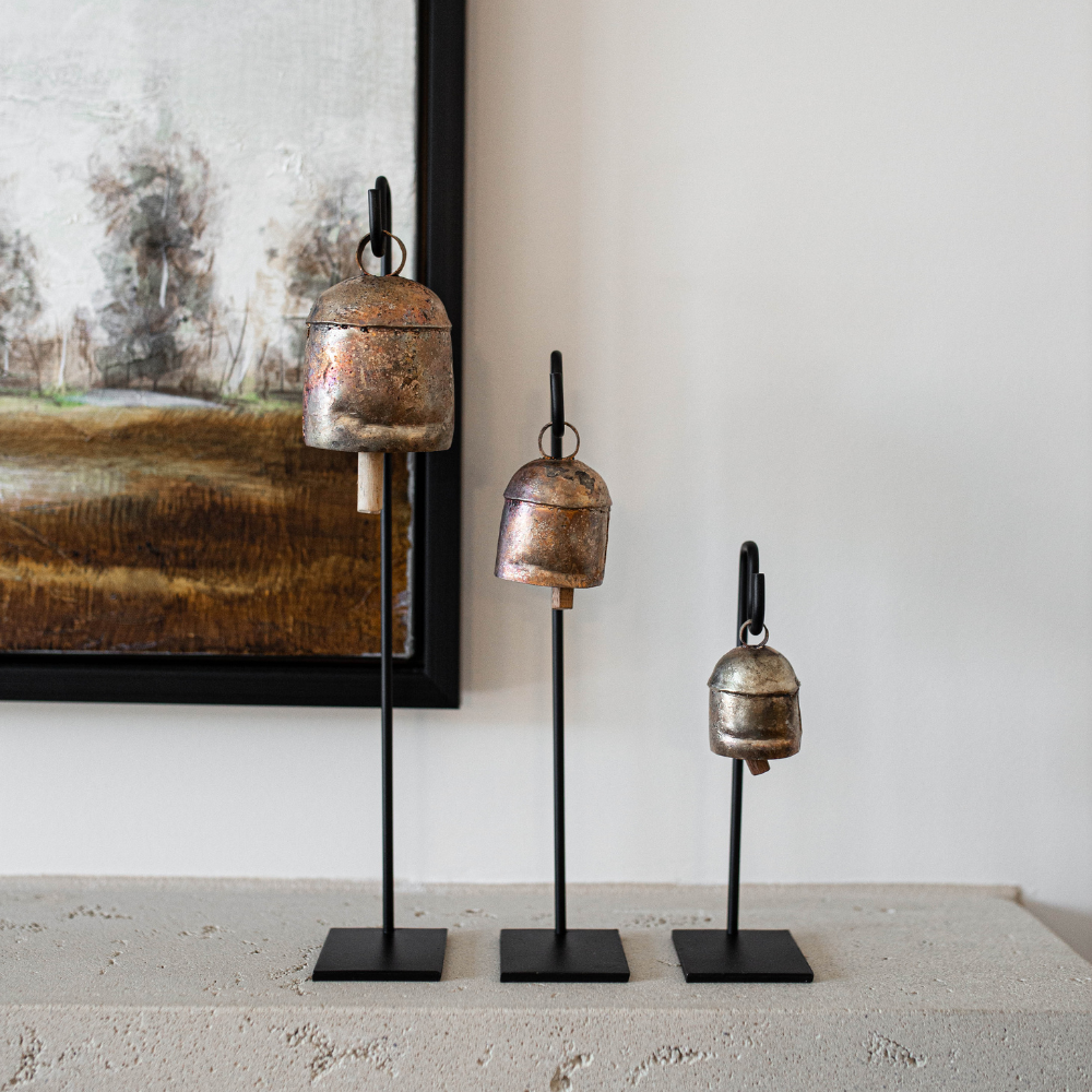 Three metallic bells on stands against a neutral wall with a painting.