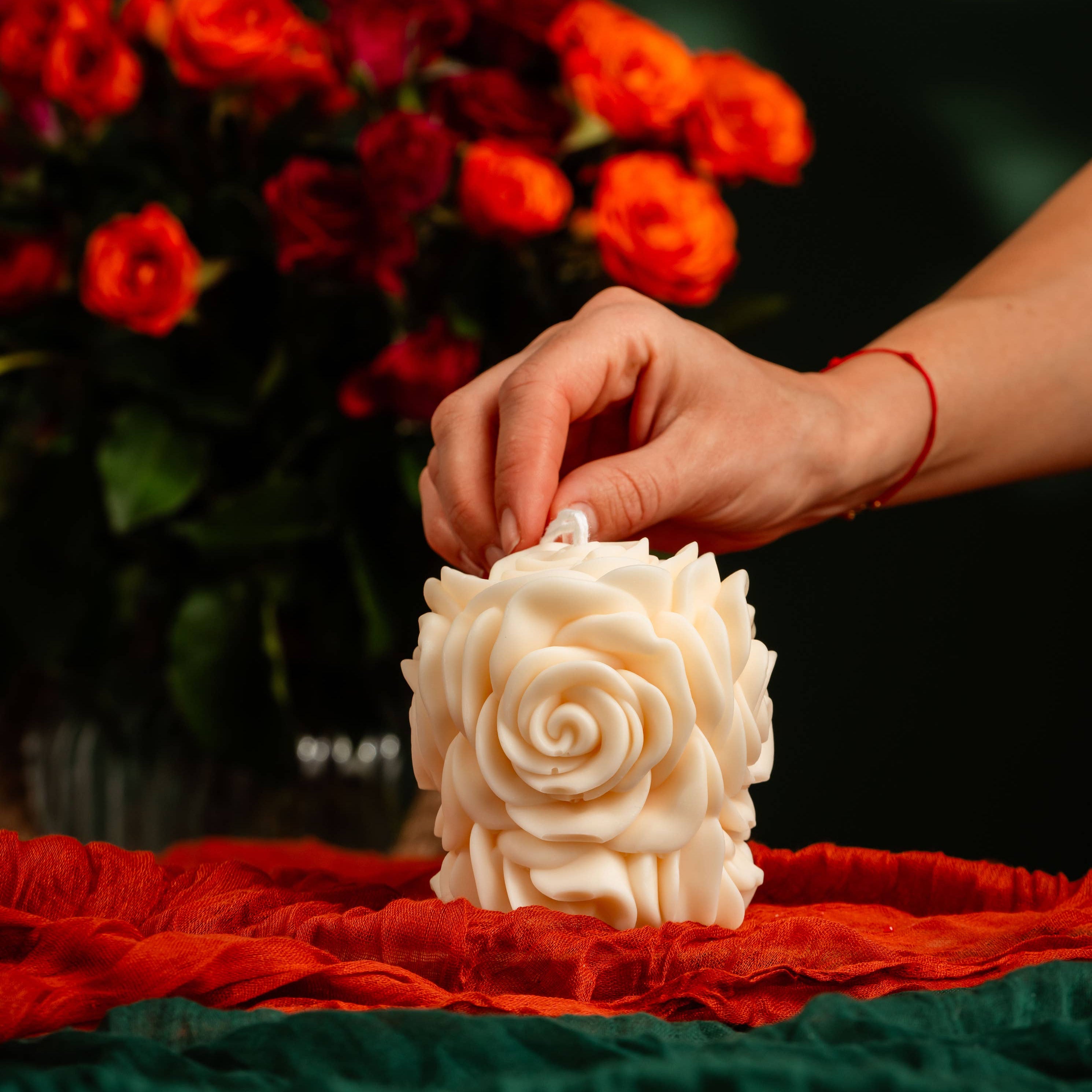 Hand placing a white rose-shaped candle on a red fabric with a blurred bouquet of red roses in the background.