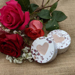 Two round bath balms with rose petals and heart shaped soap centres on a a burlap surface with red roses.