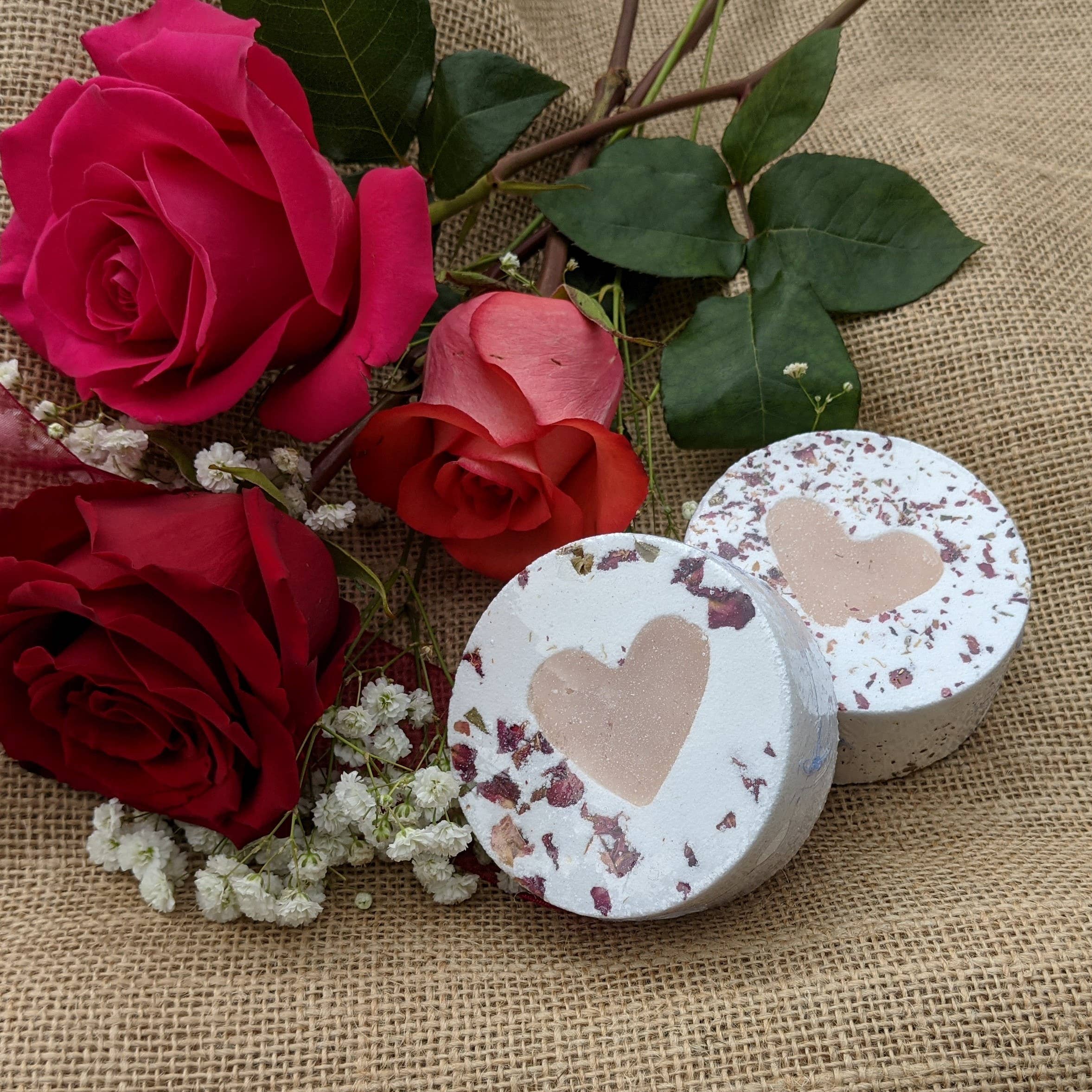 Two round bath balms with rose petals and heart shaped soap centres on a a burlap surface with red roses.