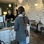 Woman wearing a floral hair clip in a gray coat standing at a counter in a cozy cafe with stone walls and wooden floors.