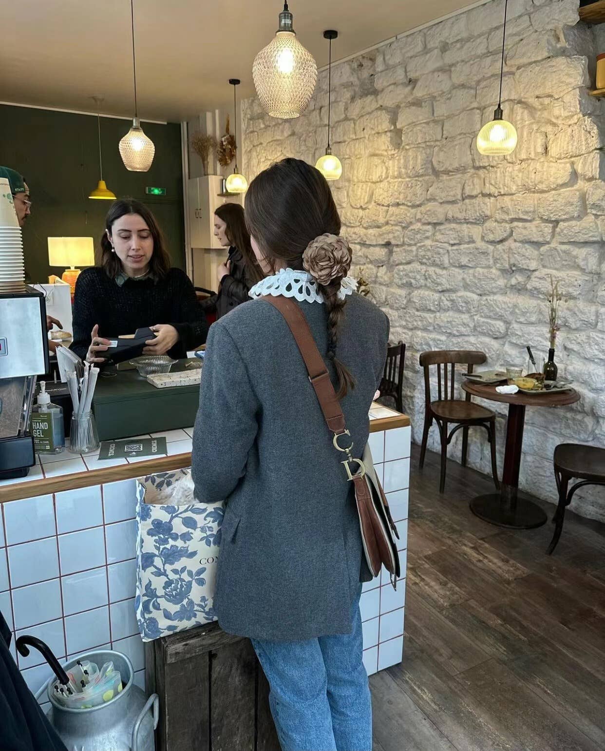 Woman wearing a floral hair clip in a gray coat standing at a counter in a cozy cafe with stone walls and wooden floors.
