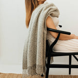 Woman sitting on a chair with a luxurious wool blend boucle throw in grey draped over her shoulders against a white wall.