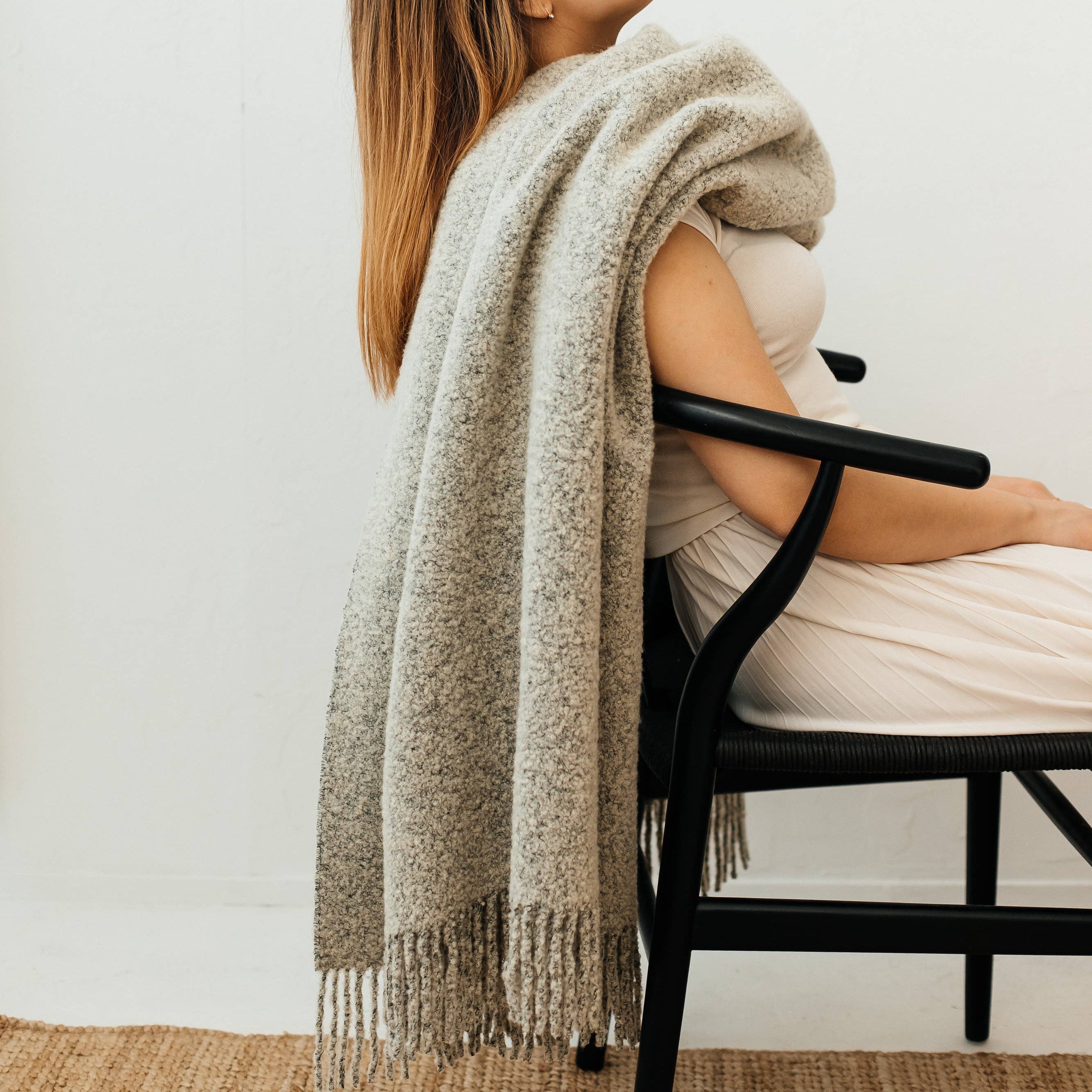 Woman sitting on a chair with a luxurious wool blend boucle throw in grey draped over her shoulders against a white wall.