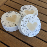 Three round bath balms with lavender petals and heart shaped soap centres on a wooden surface